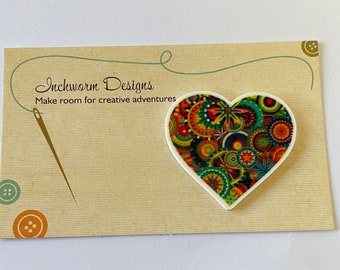 May include: A white heart-shaped pin with a colourful, abstract design of circles and swirls in shades of green, yellow, red, blue, and orange. The pin is on a brown card with the text "Inchworm Designs Make room for creative adventures".