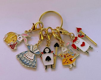 May include: A gold key chain with six charms featuring characters from Alice in Wonderland. The charms include Alice, the Queen of Hearts, the White Rabbit, the Mad Hatter, and the Cheshire Cat.