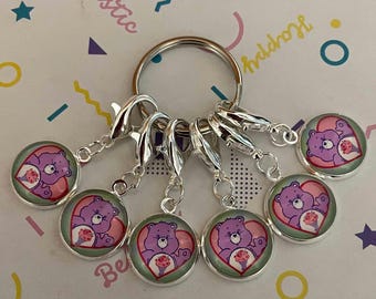 May include: A set of seven silver charm keychains with heart-shaped charms featuring purple Care Bears characters. Each charm has a different Care Bear with a different flower design. The charms are attached to silver lobster claw clasps.