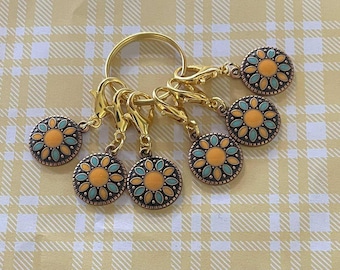 May include: A collection of six gold-coloured stitch markers with a floral design. Each marker has a round charm with an orange centre, surrounded by teal and orange petal shapes. The markers are attached to a gold ring and clasps.