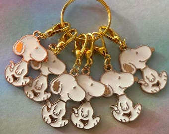 May include: A gold-toned metal keyring featuring five individual enamel charms shaped like the cartoon dog Snoopy. Each charm is white with gold outlines and details, attached to the central ring by small gold lobster claw clasps.