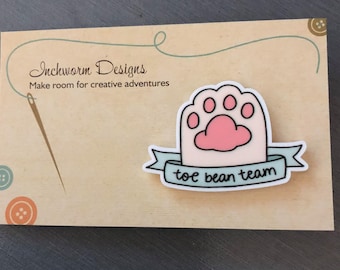 May include: A light blue and pink paw print with the text "toe bean team" on a white banner. The paw print is on a brown background with a needle and thread.