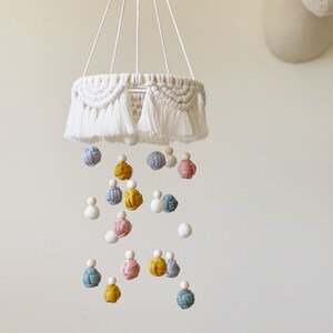 May include: A white macrame baby mobile with a circular base and multiple hanging balls in shades of blue, yellow, pink, and white.
