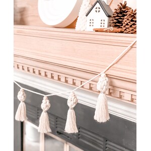 May include: A cream-colored macrame tassel garland with four tassels, each featuring a knotted design and a long, fringed bottom. The garland is strung across a mantel, with decorative items like a small house and pinecones in the background.