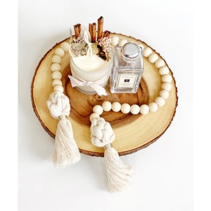 May include: A decorative arrangement featuring a wooden candle holder with a lit candle, cinnamon sticks, and dried flowers. A perfume bottle and a beaded garland with tassels are also present. The items are arranged on a wooden slice.