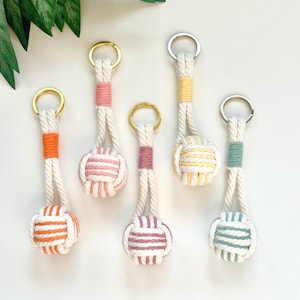 May include: Five keychains made of off-white twisted cotton rope, each featuring a spherical monkey fist knot at the bottom. The rope is accented with colored thread wraps in orange, pink, mauve, yellow, and sage green, attached to metal rings.