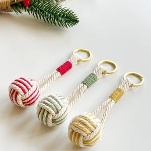 May include: Three handmade keychains with a nautical theme. Each keychain features a knotted ball design in red and white, green and white, and yellow and white. The keychains have a gold-colored ring.