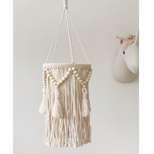 May include: A white macrame hanging lamp with wooden beads and tassels. The lamp is hanging from a white ceiling.