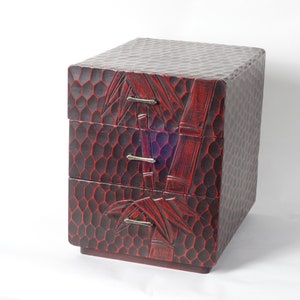 May include: A small, rectangular wooden chest with three stacked drawers. The exterior features a dark red and black finish with a textured, honeycomb-like carved pattern. A carved bamboo stalk design spans the front, accented by three silver-toned metal drawer pulls.