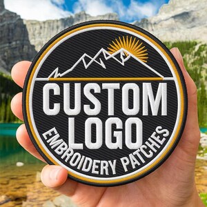 Può includere: Una toppa rotonda ricamata nera con testo bianco "CUSTOM LOGO EMBROIDERY PATCHES". Il design presenta una catena montuosa e un sole nascente in bianco e giallo, con un bordo giallo e bianco.