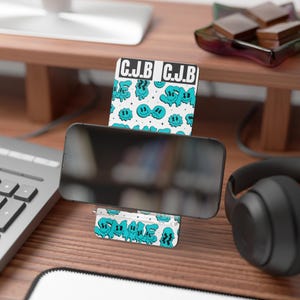 May include: A white and teal phone stand with a repeating pattern of smiling faces and the text "C.J.B." The stand is holding a black smartphone. The stand is on a wooden desk with a keyboard and headphones in the background.