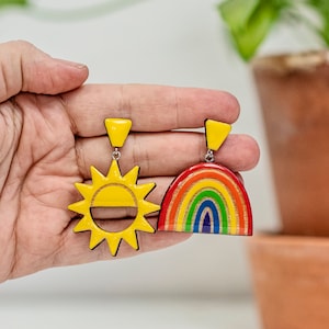 Può includere: Un paio di orecchini in legno colorati. Un orecchino ha la forma di un sole con un viso giallo e l'altro ha la forma di un arcobaleno con strisce rosse, arancioni, gialle, verdi, blu, indaco e viola.