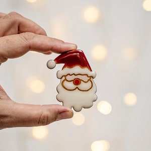 May include: A festive Santa Claus-shaped Christmas ornament. The ornament has a red hat with a white bobble, a white beard, and gold-coloured spectacles. The ornament is held between two fingers against a blurred background of lights.