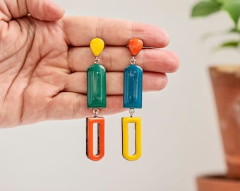 May include: A pair of long, colorful earrings with a geometric design. The earrings are made of three rectangular pieces, each a different color: green, blue, orange, and yellow. The earrings are hanging from a silver hook.