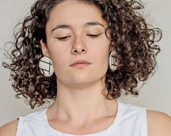 Könnte beinhalten: Eine Frau mit lockigem braunen Haar trägt weiße geometrische Ohrringe. Sie trägt ein weißes ärmelloses Oberteil.