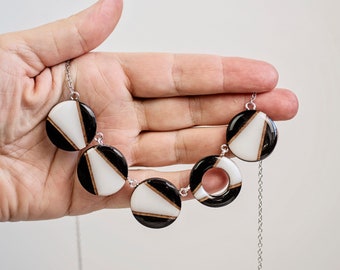 May include: A silver chain necklace with five black and white geometric resin pendant circles. The circles have a white background with black geometric shapes.