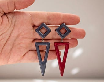 May include: A pair of geometric earrings. One earring is a dark gray diamond shape at the top with a gray triangle pendant. The other earring is a dark gray diamond shape at the top with a red triangle pendant.