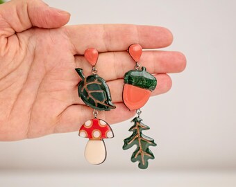 May include: A pair of quirky earrings with woodland-themed charms. One earring features a leaf and mushroom, the other an acorn and pine tree. The charms are in green, red, and peach, with a teardrop-shaped top.