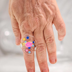 Può includere: Un anello trasparente a forma di nuvola con una pietra ovale rosa centrale incastonata in metallo dorato, circondato da piccoli punti colorati incastonati nelle tonalità del blu, giallo e rosa, creando un aspetto giocoso e strutturato.