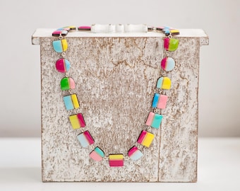 May include: A colorful necklace with a variety of shapes and colors, including pink, yellow, blue, and green. The necklace is displayed on a white wooden box. The necklace is made of multiple geometric shapes.