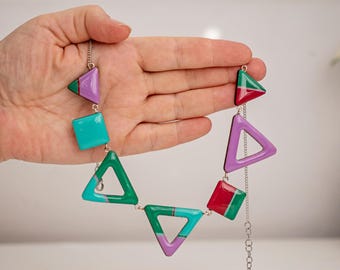 Op de afbeelding: Een kleurrijke geometrische ketting met vormen in groen, paars, rood en turquoise. De ketting bestaat uit verbonden geometrische vormen, waaronder driehoeken en vierkanten, geregen aan een zilveren ketting. De ketting wordt door een hand omhoog gehouden.