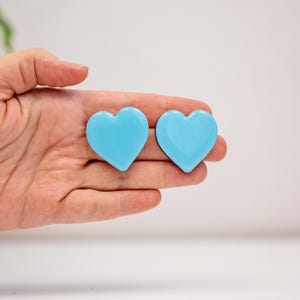 Puede incluir: Dos objetos en forma de corazón de color azul claro. Los corazones son lisos y brillantes, con una forma redondeada. Los corazones se sostienen en una mano sobre un fondo blanco. Los corazones son de un color vibrante y sólido.