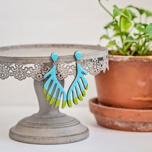 Puede incluir: Un par de pendientes colgantes de color turquesa y verde con un diseño de hojas. Los pendientes están colgados de un soporte para pasteles plateado.