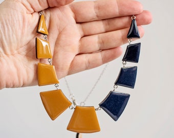 Op de afbeelding: Een statement ketting met geometrische vormen in mosterdgeel en marineblauw. De ketting is gemaakt van hars en heeft een zilveren ketting.