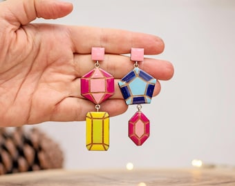 May include: A pair of colorful geometric earrings. One earring features a pink square top, a pink and fuchsia hexagon, and a yellow rectangle. The other earring has a pink square top, a blue and light blue pentagon, and a pink and fuchsia gem shape.