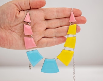 May include: A colorful necklace with geometric shapes in pink, yellow, and blue. The necklace is made of linked segments with a silver chain and a wooden accent. The design features a mix of triangular and trapezoidal shapes.