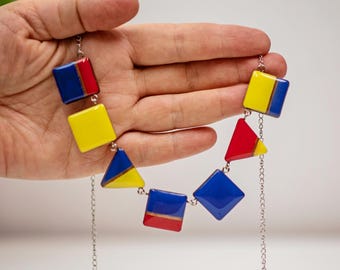 Op de afbeelding: Een kleurrijke ketting met geometrische vormen in rood, geel en blauw. De ketting heeft een zilveren ketting en bestaat uit vierkante en driehoekige hangers. Het ontwerp is modern en opvallend.