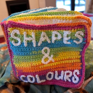 May include: A multi-coloured crocheted book with the words "Shapes & Colours" in white yarn.