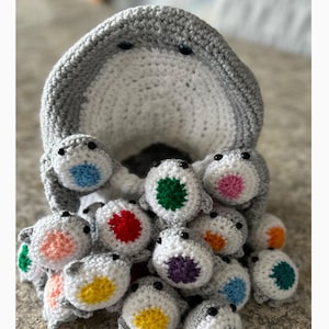 Puede incluir: Un tiburón de crochet gris con vientre blanco y ojos negros. El tiburón está sosteniendo un grupo de tiburones de crochet más pequeños con manchas de diferentes colores. Las manchas son azules, verdes, rojas, rosas, moradas, amarillas y naranjas.
