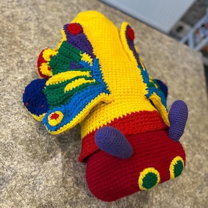May include: A crocheted caterpillar toy with a colorful butterfly design. The caterpillar is red with green eyes and purple antennas. The butterfly wings are yellow, green, blue, and purple.
