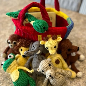 Puede incluir: Una bolsa de mano de crochet con rayas de colores, llena de juguetes de animales de crochet, incluyendo una serpiente, un mono, un león, un elefante, una jirafa, una rana y un oso.