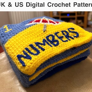 Puede incluir: Una pila de cuadrados de crochet en azul y amarillo con la palabra "NUMBERS" bordada en azul en el cuadrado amarillo. El cuadrado superior tiene una sombrilla de crochet roja, amarilla y blanca. El número "1" está bordado en el cuadrado azul.