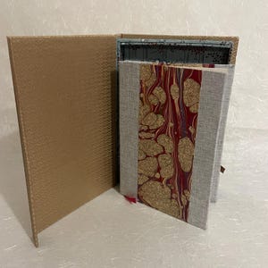 May include: An open, gold-colored box with a textured pattern, revealing a book with a marbled design in red, gold, and gray. The book has a gray linen cover and a red ribbon bookmark.