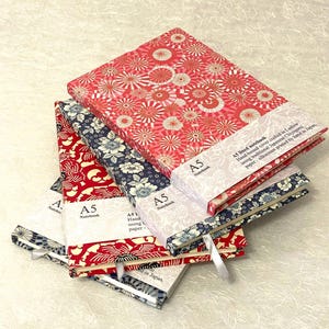 Könnte beinhalten: Eine Sammlung von A5-Notizbüchern mit gemusterten Einbänden. Die Notizbücher zeigen florale und geometrische Designs in Rot, Blau und Weiß. Jedes Notizbuch hat ein Leseband und ein Papierband mit Text. Die Notizbücher sind handgebunden und in Japan hergestellt.