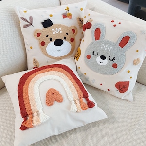 Könnte beinhalten: Drei dekorative Kissen mit gestickten Designs. Ein Kissen zeigt einen Regenbogen, ein anderes einen Bären und das dritte einen Hasen. Die Kissen liegen auf einem hellbeigen Sofa.