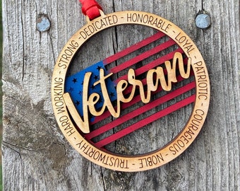 May include: Wooden ornament with a red, white, and blue American flag design. The word "Veteran" is written in a cursive font over the flag. The ornament is surrounded by a circle of text that reads "Strong - Dedicated - Honorable - Loyal - Patriotic - Courageous - Noble - Trustworthy - Hard Working".
