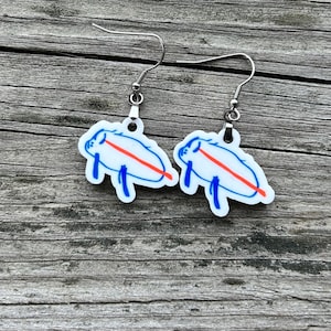 May include: A pair of dangle earrings featuring a white design with a blue outline and a red stripe. The earrings have a silver-colored hook. The design appears to be a stylized animal.