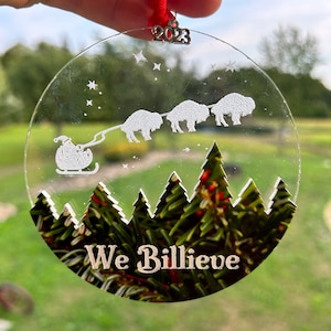 May include: A clear glass Christmas ornament with a white silhouette of Santa Claus pulling a sleigh with four buffalo. The ornament has a green pine tree forest design at the bottom and the text "We Billieve" in white. The ornament has a silver 2023 charm attached to the top.