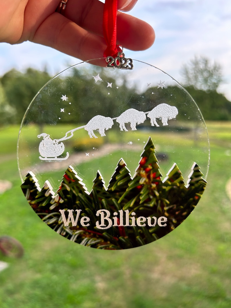 May include: A clear glass Christmas ornament with a white silhouette of Santa Claus pulling a sleigh with four buffalo. The ornament has a green pine tree forest design at the bottom and the text "We Billieve" in white. The ornament has a silver 2023 charm attached to the top.