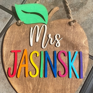 May include: A wooden apple-shaped sign with a green leaf on top. The sign says "Mrs. Jasinski" in rainbow colours.
