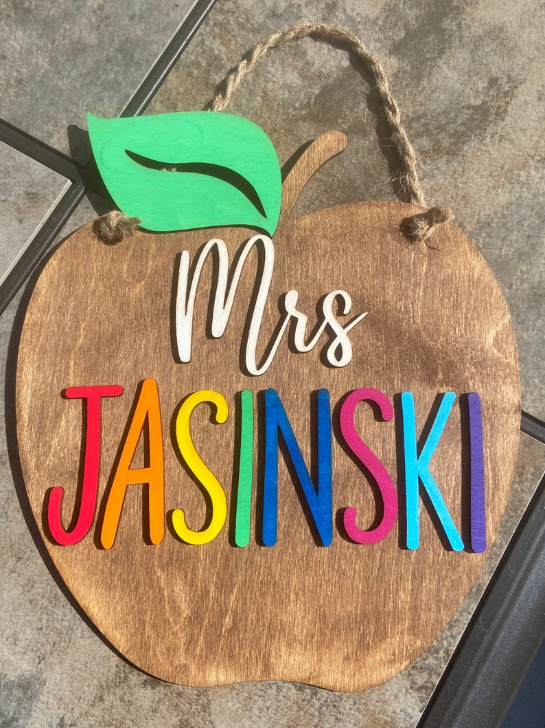 May include: A wooden apple-shaped sign with a green leaf on top. The sign says "Mrs. Jasinski" in rainbow colours.