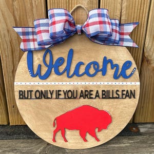 May include: A round wooden door hanger with a red buffalo silhouette, a blue welcome sign, and a red and blue plaid bow. The sign reads "Welcome, but only if you are a Bills fan."