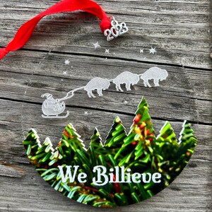 May include: A round, clear glass ornament with a white silhouette of Santa Claus in a sleigh pulled by four buffalo. The ornament has a green and brown image of a forest with the text "We Billieve" in white. The ornament has a silver 2023 charm attached.