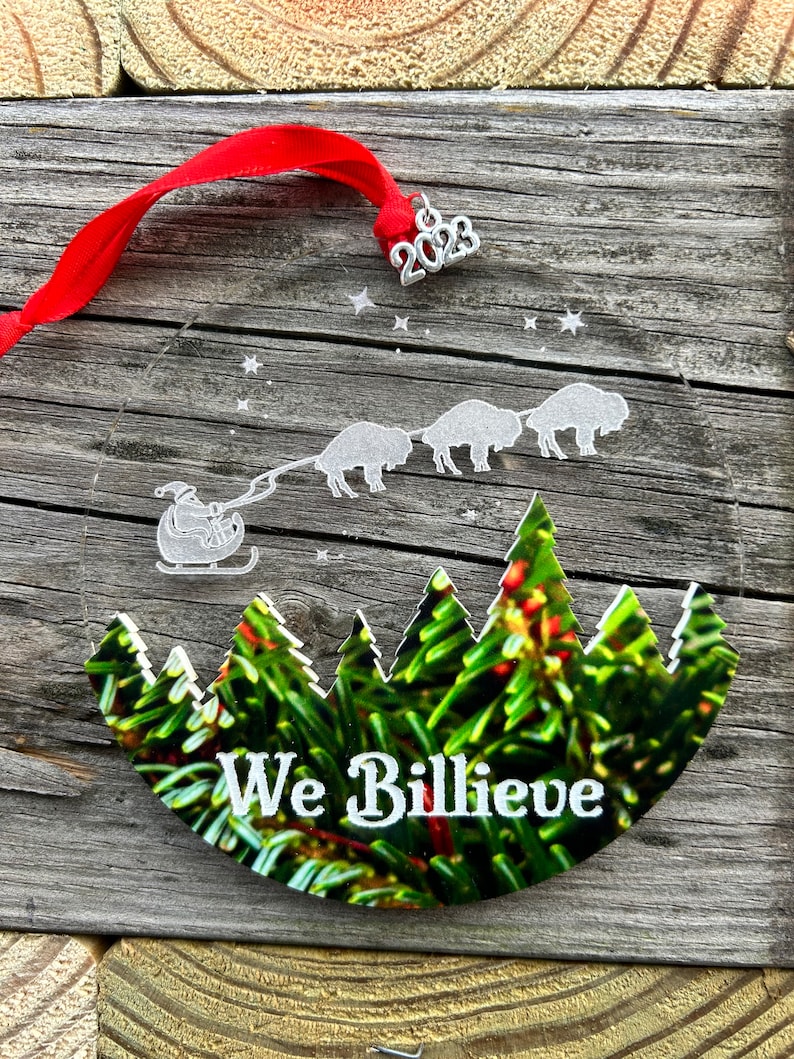 May include: A round, clear glass ornament with a white silhouette of Santa Claus in a sleigh pulled by four buffalo. The ornament has a green and brown image of a forest with the text "We Billieve" in white. The ornament has a silver 2023 charm attached.