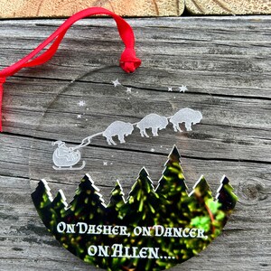 May include: A round, clear glass ornament with a white silhouette of a sleigh pulled by four buffalo. The ornament has a red ribbon attached and the text "On Dasher, On Dancer, On Allen..." is printed on the bottom.