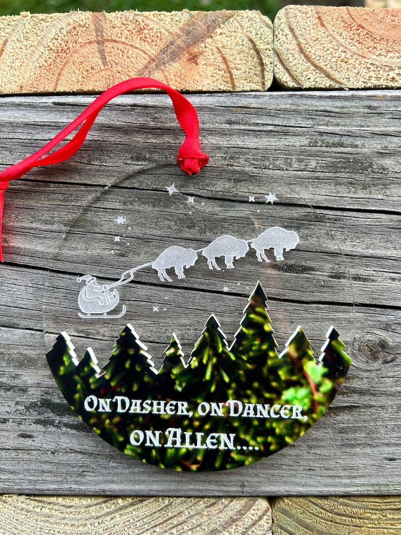 May include: A round, clear glass ornament with a white silhouette of a sleigh pulled by four buffalo. The ornament has a red ribbon attached and the text "On Dasher, On Dancer, On Allen..." is printed on the bottom.